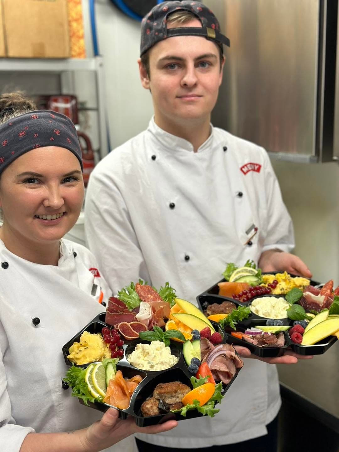 To ansatte holder fram koldtbordesker med spekemat, roastbiff, eggerøre, potetsalat og frukt.